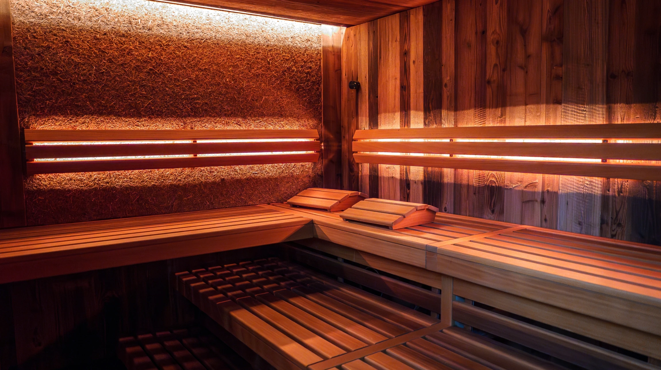 Intérieur d'un sauna infrarouge moderne avec parois en bois clair et rangées de panneaux infrarouges discrets visibles. Un ou deux clients assis confortablement sur les banquettes, dans une atmosphère chaleureuse et apaisante. La lumière douce et homogène émise par les panneaux infrarouges crée une ambiance relaxante. Le design est épuré et contemporain.